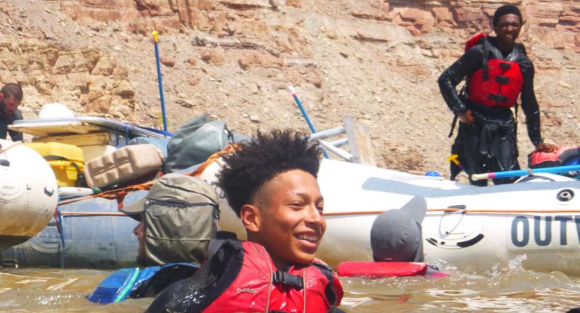 A person wearing a life jacket smiles in chest-deep water. Behind them are rafts with other people and tall canyon walls. 